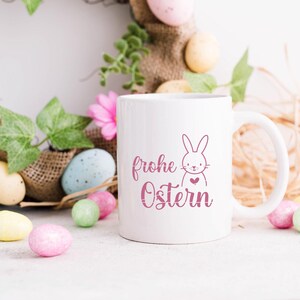 K&ouml;nnte beinhalten: Wei&szlig;e Keramiktasse mit einem rosa Hasen und dem Text "frohe Ostern" in rosa Schrift. Die Tasse ist umgeben von pastellfarbenen Ostereiern und einem Jutekranz.