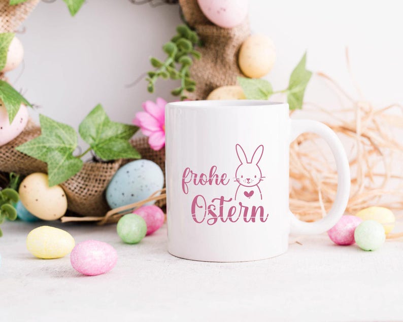 K&ouml;nnte beinhalten: Wei&szlig;e Keramiktasse mit einem rosa Hasen und dem Text "frohe Ostern" in rosa Schrift. Die Tasse ist umgeben von pastellfarbenen Ostereiern und einem Jutekranz.