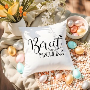 May include: White pillow with black text that reads "Bereit f&uuml;r den Fr&uuml;hling" which translates to "Ready for Spring". The pillow is surrounded by white and yellow Easter eggs.
