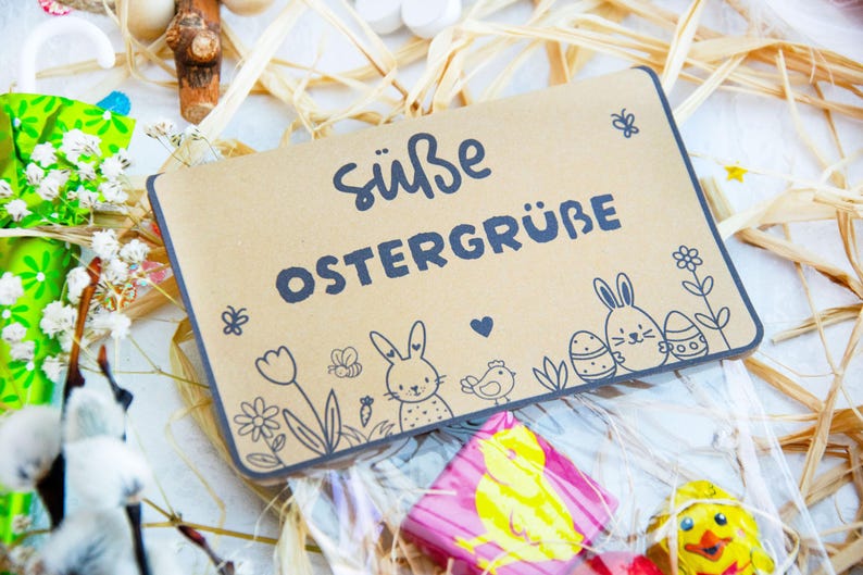 May include: A brown paper bag with a cute Easter greeting that says "S&uuml;&szlig;e Ostergr&uuml;&szlig;e" and features a bunny, flowers, and eggs. The bag is filled with chocolate Easter treats.