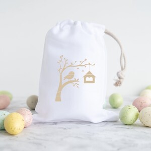 May include: White cotton drawstring bag with a gold silhouette of a bird on a branch with a birdhouse. The bag is surrounded by pastel colored speckled Easter eggs.