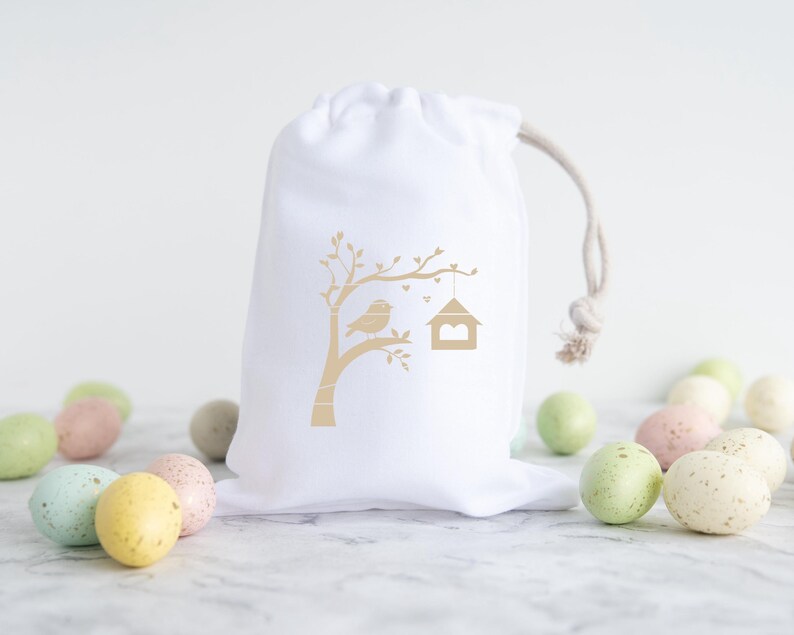 May include: White cotton drawstring bag with a gold silhouette of a bird on a branch with a birdhouse. The bag is surrounded by pastel colored speckled Easter eggs.