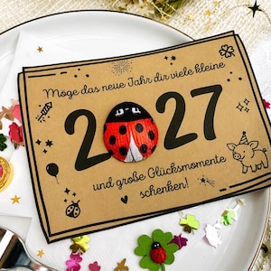 May include: A rectangular tan card featuring the black text "M&ouml;ge das neue Jahr dir viele kleine 2027 und gro&szlig;e Gl&uuml;cksmomente schenken!" with a red and black ladybug attached to the zero, alongside illustrations of a pig, stars, and fireworks.