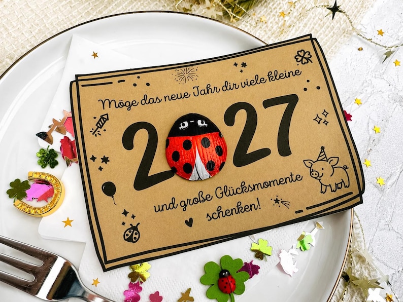 May include: A rectangular tan card featuring the black text "M&ouml;ge das neue Jahr dir viele kleine 2027 und gro&szlig;e Gl&uuml;cksmomente schenken!" with a red and black ladybug attached to the zero, alongside illustrations of a pig, stars, and fireworks.