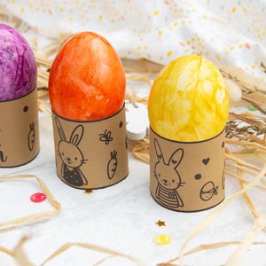 May include: Four Easter eggs decorated with different colors and patterns are displayed in brown cardboard holders with bunny illustrations. The holders have the word "ern" written on one of them.