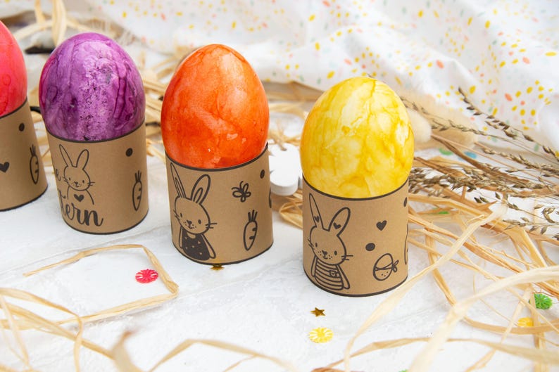 May include: Four Easter eggs decorated with different colors and patterns are displayed in brown cardboard holders with bunny illustrations. The holders have the word "ern" written on one of them.