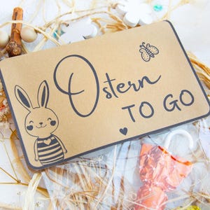 Op de afbeelding: Een bruine papieren cadeauzak met een konijnenillustratie en de tekst "Ostern TO GO". De zak is gevuld met kleine verpakte snoepjes en is omgeven door stro en andere paasversieringen.