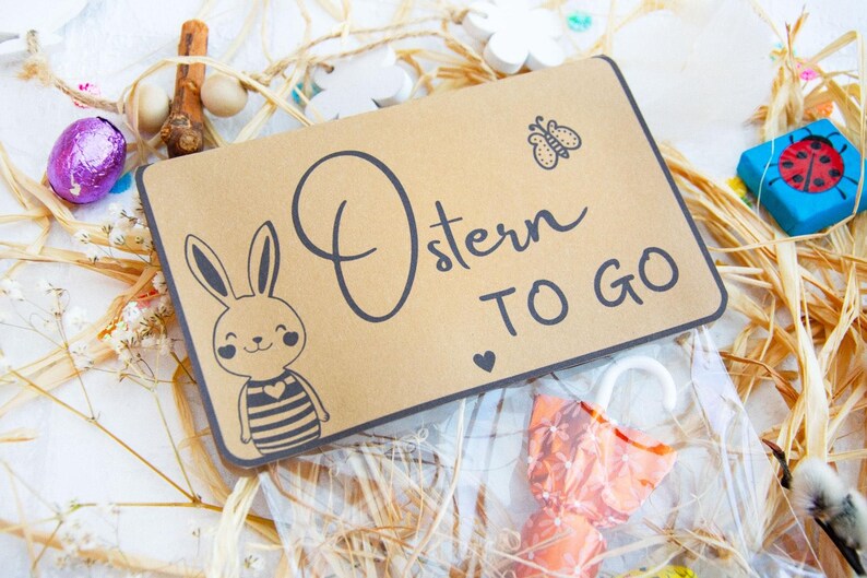 Op de afbeelding: Een bruine papieren cadeauzak met een konijnenillustratie en de tekst "Ostern TO GO". De zak is gevuld met kleine verpakte snoepjes en is omgeven door stro en andere paasversieringen.