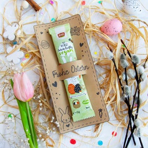 May include: A brown card with the text "Frohe Ostern" (Happy Easter) and a bunny rabbit illustration. A green and white chocolate bar with the text "Die Echte Schokolade Vegan" (The Real Chocolate Vegan) is on the card. The card is surrounded by straw, white flowers, and pastel-colored Easter eggs.