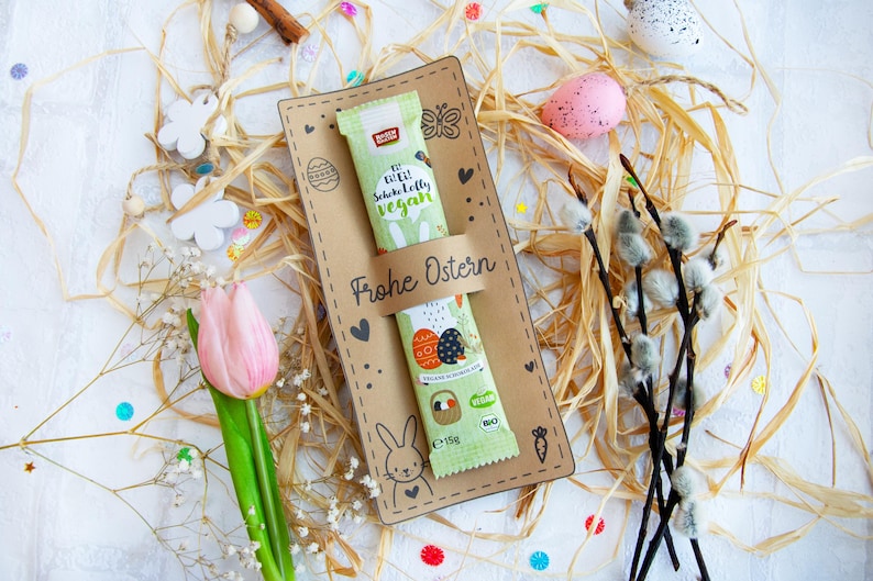 May include: A brown card with the text "Frohe Ostern" (Happy Easter) and a bunny rabbit illustration. A green and white chocolate bar with the text "Die Echte Schokolade Vegan" (The Real Chocolate Vegan) is on the card. The card is surrounded by straw, white flowers, and pastel-colored Easter eggs.