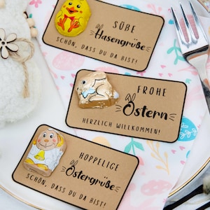 May include: An Easter-themed display on a white plate. Features three kraft paper cards with German text and chocolate candies shaped like a chick, a bunny, and a lamb. A plush bunny and a silver fork are also present.