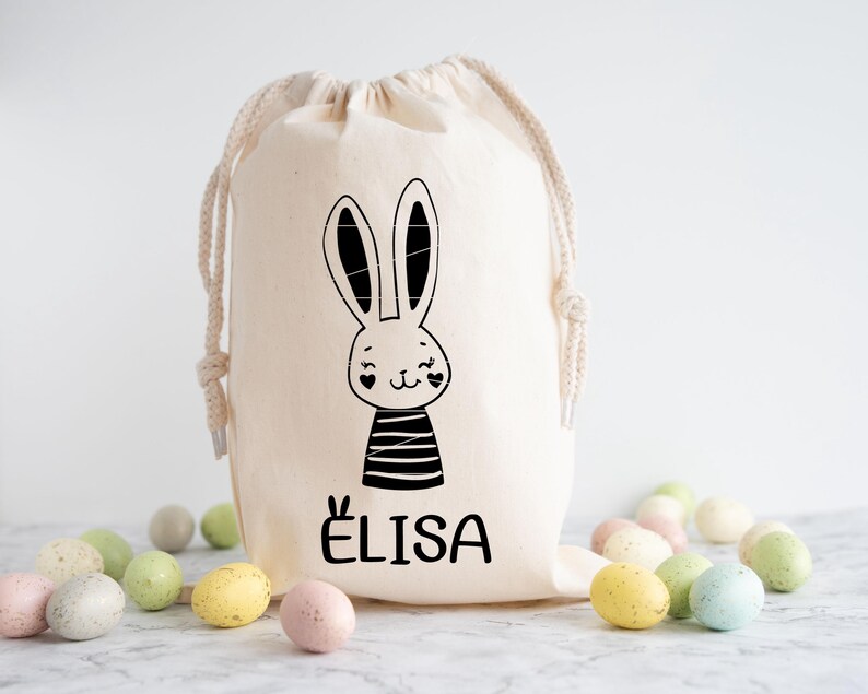 Op de afbeelding: Een witte canvas trekkoordtas met een zwart-witte konijnenillustratie en de naam "ELISA" gedrukt op de voorkant. De tas is gevuld met pastelkleurige paaseieren.