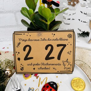 May include: A rectangular tan card with black text reading "M&ouml;ge das neue Jahr dir viele kleine 2 27 und gro&szlig;e Gl&uuml;cksmomente schenken!" featuring illustrations of a firework, stars, a balloon, a ladybug, and a pig wearing a party hat.