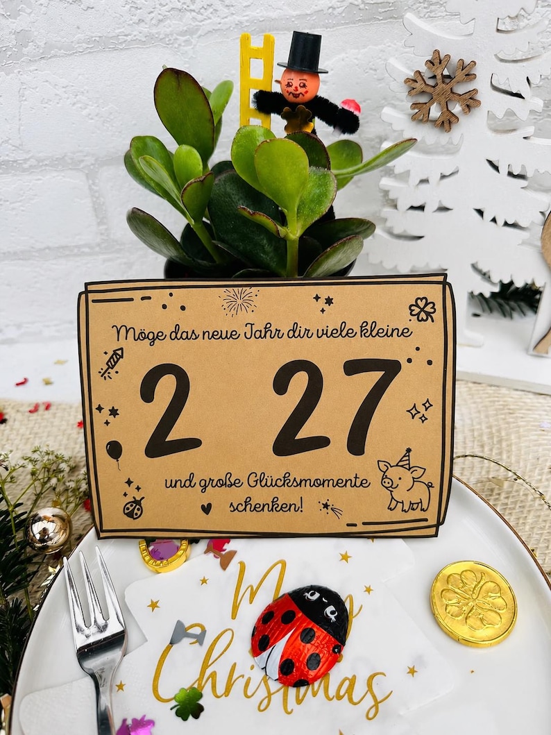 May include: A rectangular tan card with black text reading "M&ouml;ge das neue Jahr dir viele kleine 2 27 und gro&szlig;e Gl&uuml;cksmomente schenken!" featuring illustrations of a firework, stars, a balloon, a ladybug, and a pig wearing a party hat.