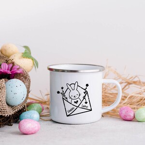 May include: White enamel mug with a black and white illustration of a bunny in an envelope. The envelope has the text "Hassen" and "Liebe" on it. The mug is surrounded by pastel colored Easter eggs and straw.