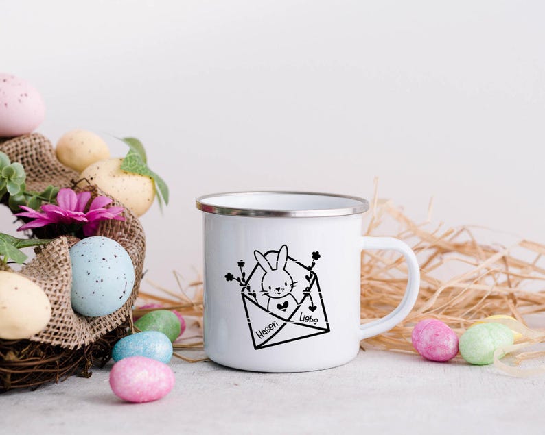 May include: White enamel mug with a black and white illustration of a bunny in an envelope. The envelope has the text "Hassen" and "Liebe" on it. The mug is surrounded by pastel colored Easter eggs and straw.