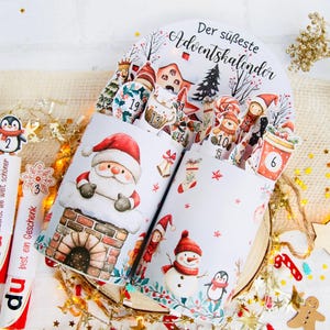 Op de afbeelding: Een adventskalender met illustraties van de kerstman, een sneeuwpop en een pinguïn. De kalender is versierd met kerstafbeeldingen en de tekst "Der süßeste Adventskalender". Twee chocoladerepen met pinguïn-toppers zijn ook zichtbaar.