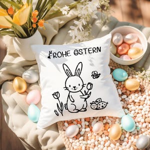 May include: A white square cushion featuring a black bunny illustration holding a flower, with the words "FROHE OSTERN" above. The cushion is surrounded by pastel-coloured Easter eggs and a small bowl of eggs.