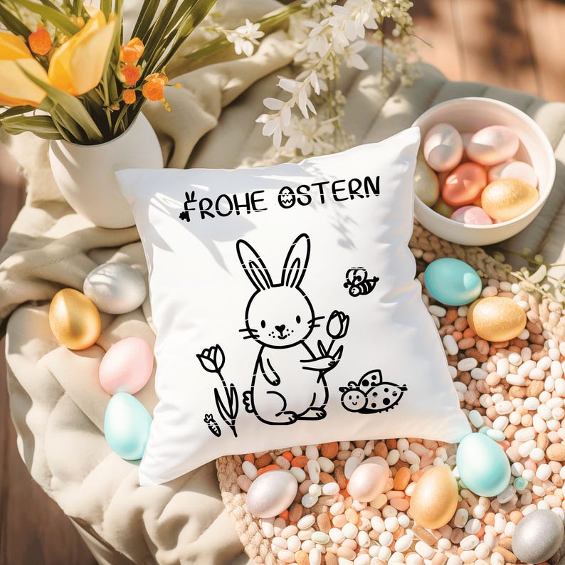 May include: A white square cushion featuring a black bunny illustration holding a flower, with the words "FROHE OSTERN" above. The cushion is surrounded by pastel-coloured Easter eggs and a small bowl of eggs.