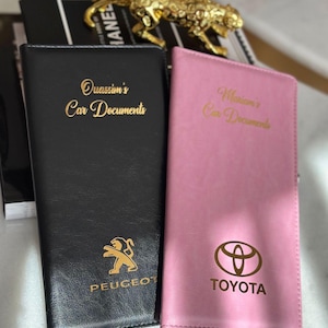 May include: Black and pink car document holders. The black holder has "Ouassim's Car Documents" in gold lettering and a gold Peugeot logo. The pink holder has "Mariam's Car Documents" and a gold Toyota logo.