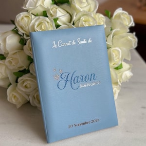 May include: A light blue health record book with the name "Haron" in glittery blue script. The text "Le Carnet de Sant&eacute; de" and the date "20 Novembre 2024" are also visible. A bouquet of white roses is in the background.