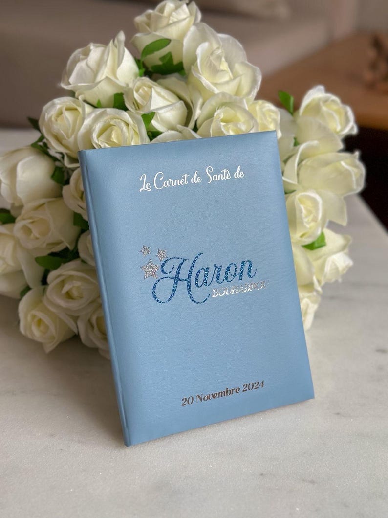 May include: A light blue health record book with the name "Haron" in glittery blue script. The text "Le Carnet de Sant&eacute; de" and the date "20 Novembre 2024" are also visible. A bouquet of white roses is in the background.