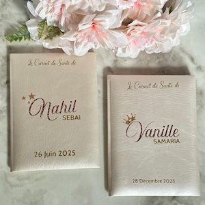 May include: Two off-white health record books with gold lettering and dates. The top of each book reads "Le Carnet de Sant&eacute; de". One book is labeled "Nahil SEBAI 26 Juin 2025". The other book is labeled "Vanille SAMARIA 28 D&eacute;cembre 2025".