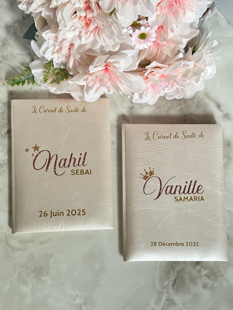 May include: Two off-white health record books with gold lettering and dates. The top of each book reads "Le Carnet de Sant&eacute; de". One book is labeled "Nahil SEBAI 26 Juin 2025". The other book is labeled "Vanille SAMARIA 28 D&eacute;cembre 2025".