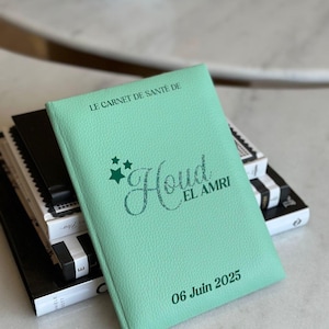 May include: A light green health record book with the text "LE CARNET DE SANT&Eacute; DE" at the top. The name "Houd EL AMRI" is written in a cursive font, with the date "06 Juin 2025" at the bottom. The book is stacked on top of other books.