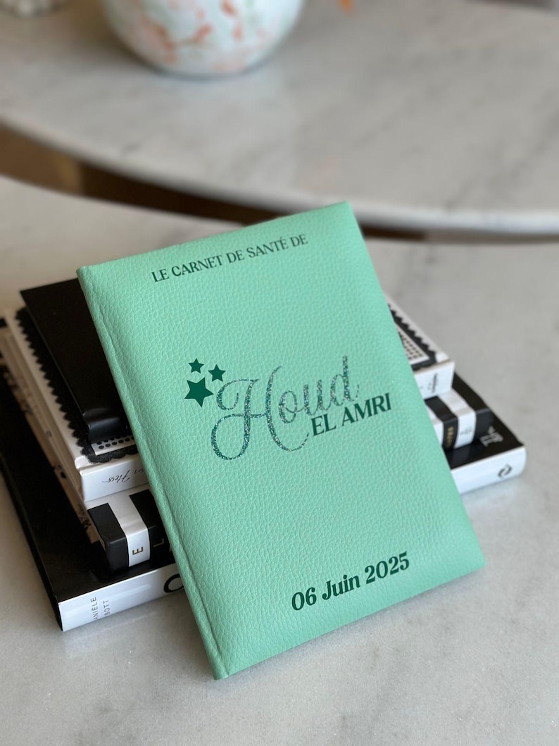 May include: A light green health record book with the text "LE CARNET DE SANT&Eacute; DE" at the top. The name "Houd EL AMRI" is written in a cursive font, with the date "06 Juin 2025" at the bottom. The book is stacked on top of other books.