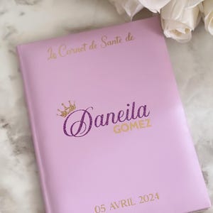 May include: A light pink notebook with gold lettering and a gold and purple glitter crown. The text on the cover reads "Le Carnet de Sante de Daneila Gomez 05 Avril 2024". White roses are in the background.
