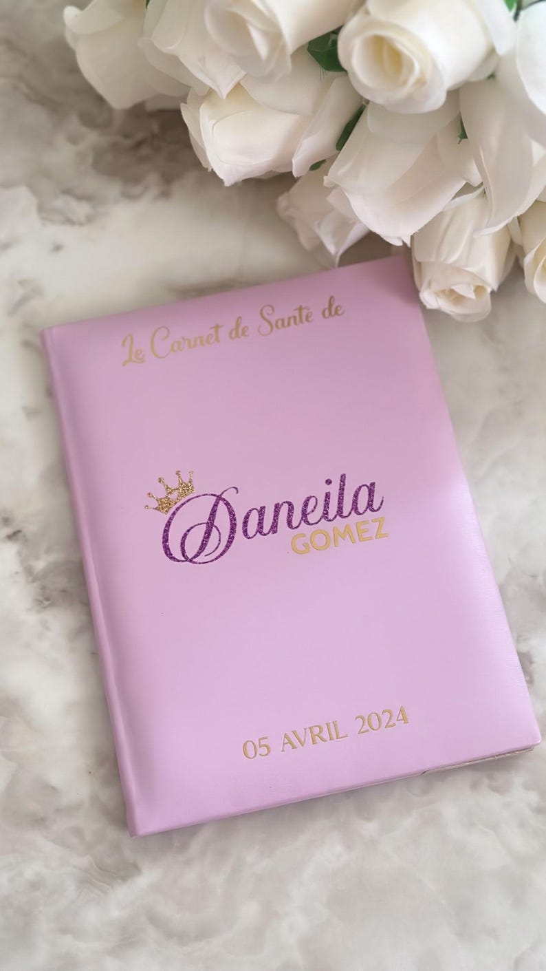 May include: A light pink notebook with gold lettering and a gold and purple glitter crown. The text on the cover reads "Le Carnet de Sante de Daneila Gomez 05 Avril 2024". White roses are in the background.