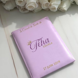 May include: A lavender-colored notebook with gold lettering and the name "T&eacute;ha Courla" in a glittery purple font. The text "27 JUIN 2019" is also printed on the cover. White roses are in the background.