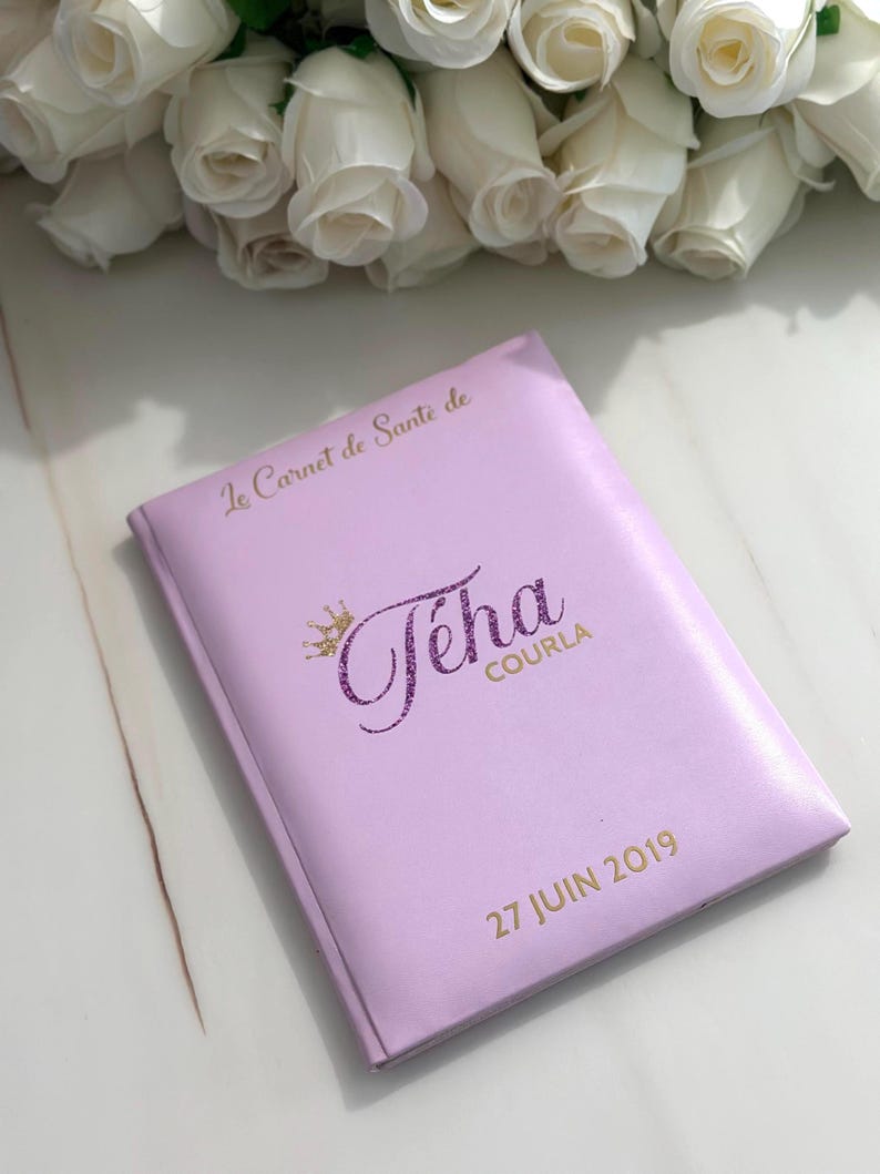 May include: A lavender-colored notebook with gold lettering and the name "T&eacute;ha Courla" in a glittery purple font. The text "27 JUIN 2019" is also printed on the cover. White roses are in the background.