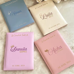 May include: Four personalized baby health record books in pastel colors: light blue, cream, and two shades of pink. Each book features a name and date in gold or glittery script. White roses are in the background.