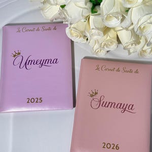 May include: Two personalized notebooks, one lavender and one pink, with gold lettering and a crown design. The lavender notebook is labeled "Umeyma" and the year "2025." The pink notebook is labeled "Sumaya" and the year "2026."