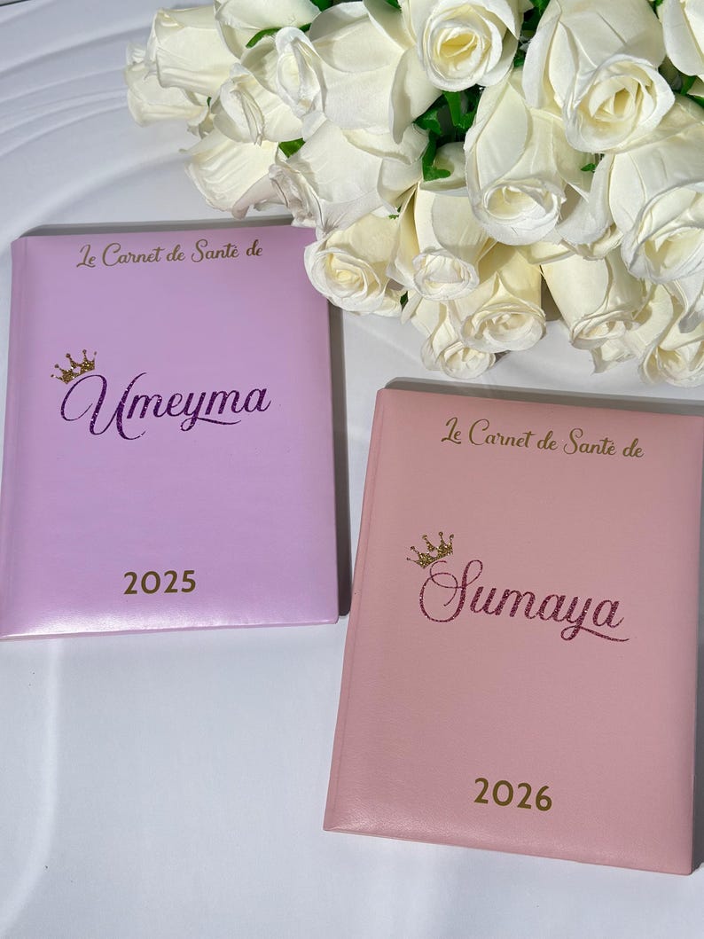 May include: Two personalized notebooks, one lavender and one pink, with gold lettering and a crown design. The lavender notebook is labeled "Umeyma" and the year "2025." The pink notebook is labeled "Sumaya" and the year "2026."