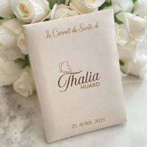 May include: A cream-colored book with the text "Le Carnet de Sante de" and the name "Thalia Huard" in a decorative font. The date "25 Avril 2025" is printed at the bottom. White roses are in the background.