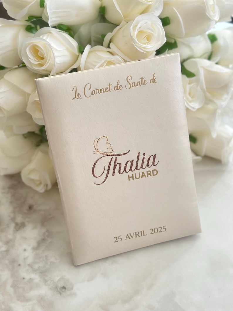 May include: A cream-colored book with the text "Le Carnet de Sante de" and the name "Thalia Huard" in a decorative font. The date "25 Avril 2025" is printed at the bottom. White roses are in the background.