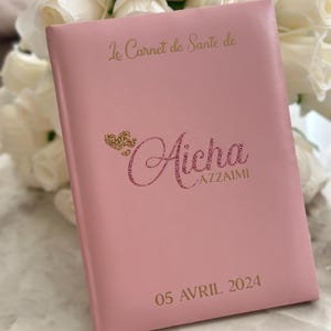 May include: A pink health record book with gold lettering and a glittery name. The cover reads "Le Carnet de Sant&eacute; de" and "Aicha AZZAIMI" with the date "05 AVRIL 2024". White roses are in the background.