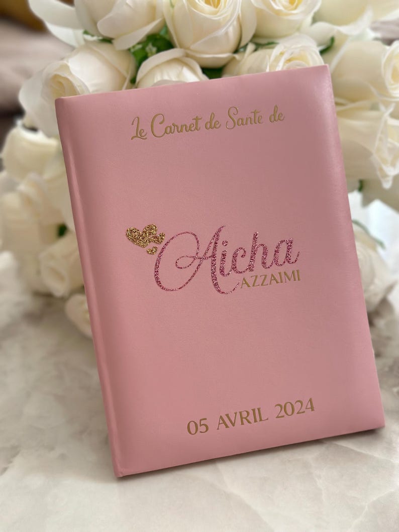 May include: A pink health record book with gold lettering and a glittery name. The cover reads "Le Carnet de Sant&eacute; de" and "Aicha AZZAIMI" with the date "05 AVRIL 2024". White roses are in the background.