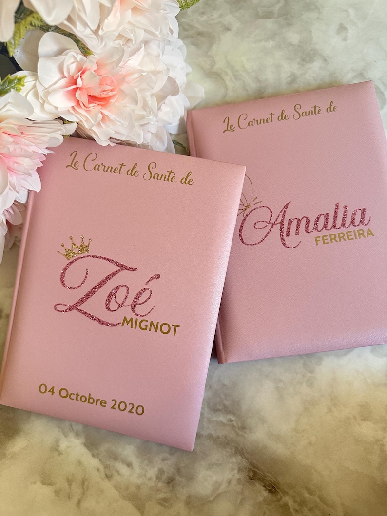 May include: Two pink health record books with gold lettering and glitter accents. One book is personalized with the name "Zo&eacute; MIGNOT" and the date "04 Octobre 2020". The other book is personalized with the name "Amalia FERREIRA".
