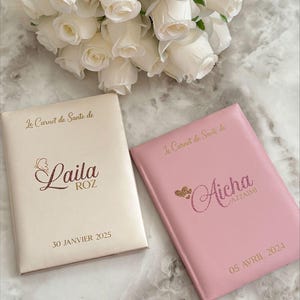 May include: Two personalized health record books, one cream and one pink, with gold lettering and glitter accents. The cream book says "Laila" and the pink book says "Aicha". A bouquet of white roses is in the background.