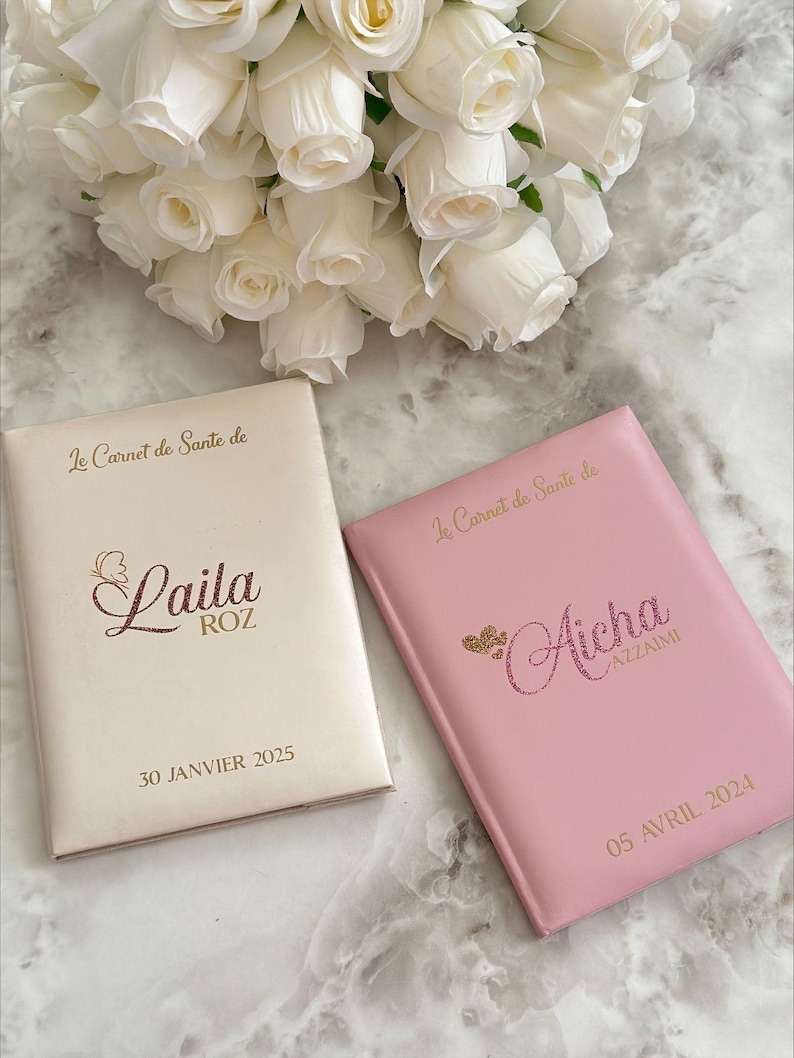 May include: Two personalized health record books, one cream and one pink, with gold lettering and glitter accents. The cream book says "Laila" and the pink book says "Aicha". A bouquet of white roses is in the background.