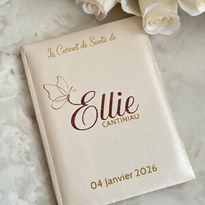 May include: A cream-colored book with gold text and a butterfly design. The cover reads "Le Carnet de Sante de Ellie CANTINIAU 04 Janvier 2026". The book is likely a health record or journal, with a personalized name.