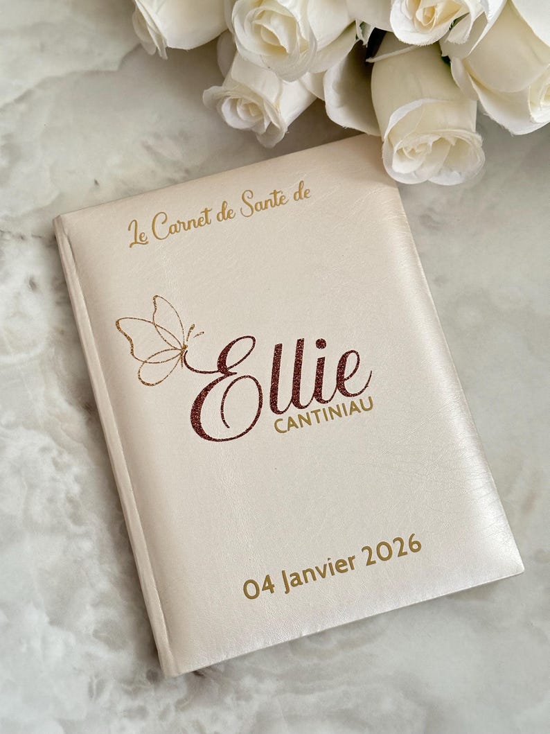 May include: A cream-colored book with gold text and a butterfly design. The cover reads "Le Carnet de Sante de Ellie CANTINIAU 04 Janvier 2026". The book is likely a health record or journal, with a personalized name.