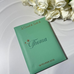 May include: A mint green journal with the text "Le Carnet de Sante de Thoma" in gold lettering. The date "18 FEVRIER 2025" is also printed on the journal. A bouquet of white roses is in the background.
