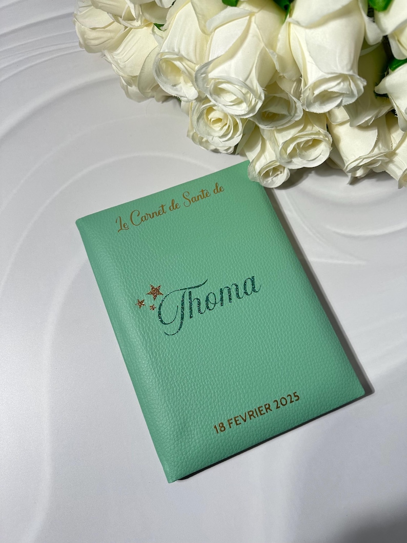 May include: A mint green journal with the text "Le Carnet de Sante de Thoma" in gold lettering. The date "18 FEVRIER 2025" is also printed on the journal. A bouquet of white roses is in the background.