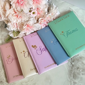 May include: A collection of colorful notebooks, each with a unique name and date, arranged near a bouquet of pink and white flowers. The notebooks are in shades of pink, cream, lavender, blue, and mint green, with gold lettering.