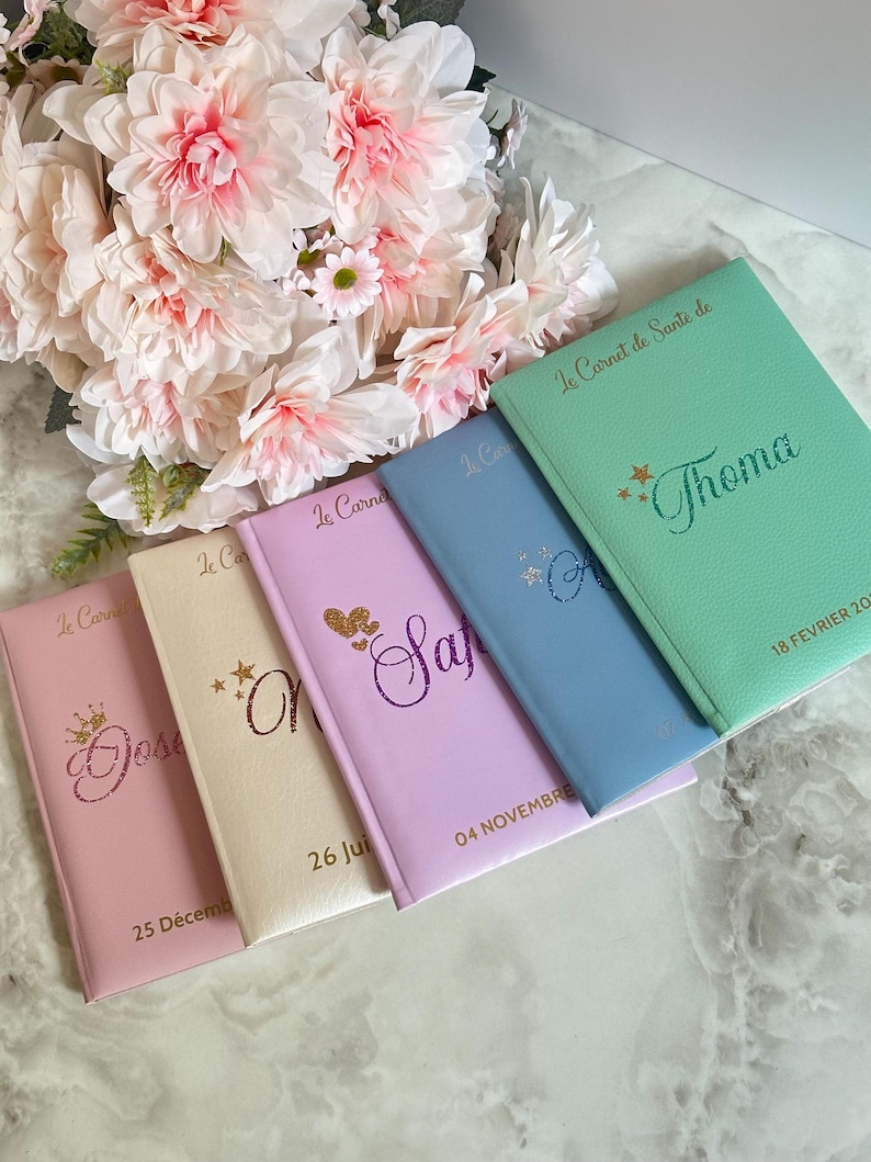 May include: A collection of colorful notebooks, each with a unique name and date, arranged near a bouquet of pink and white flowers. The notebooks are in shades of pink, cream, lavender, blue, and mint green, with gold lettering.